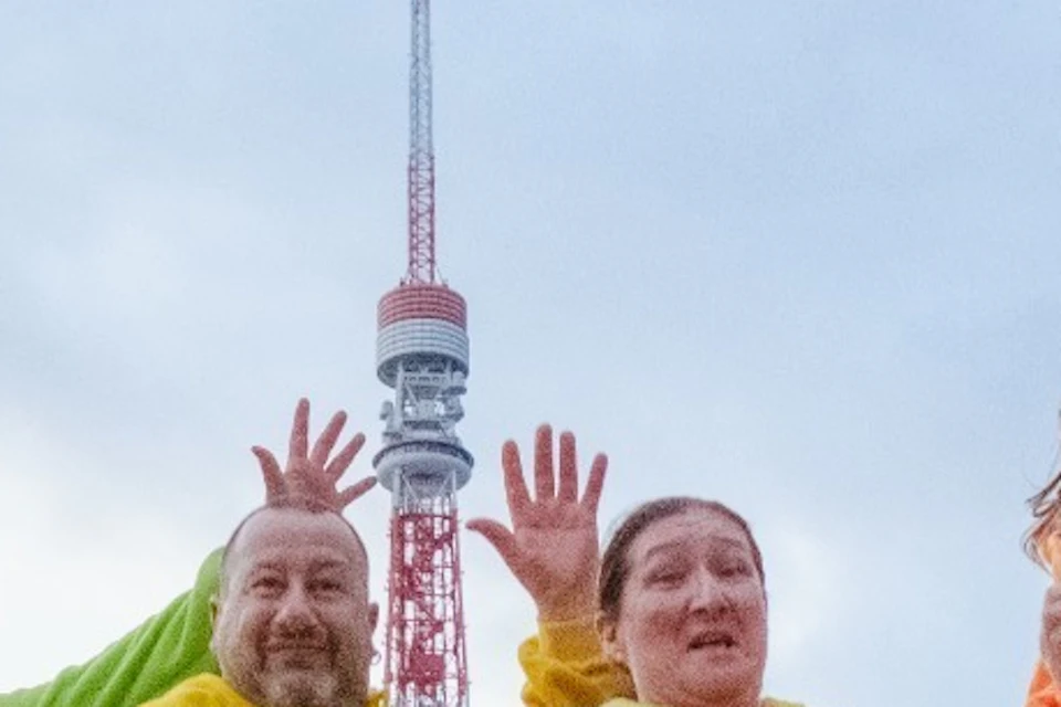 A group of people in colorful costumes joyfully jumping outdoors near a tower and trees.