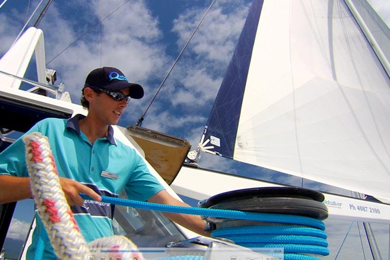 A person in a teal shirt and black cap operates ropes on a sailboat with sails unfurled under a partly cloudy sky.