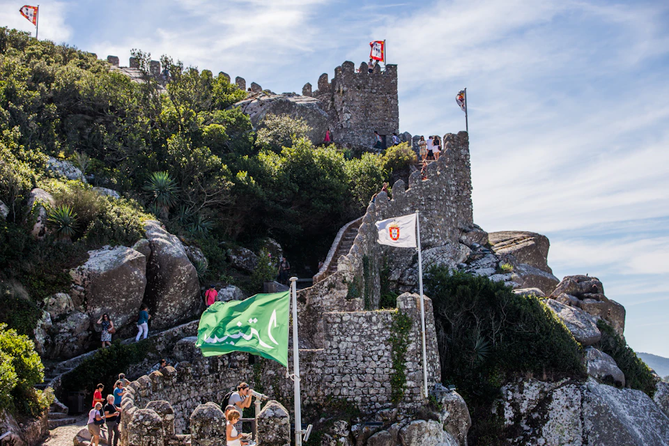 Stone castle ruins atop a hill with flags and tourists scattered around. Dense green foliage surrounds the structure under a bright sky.