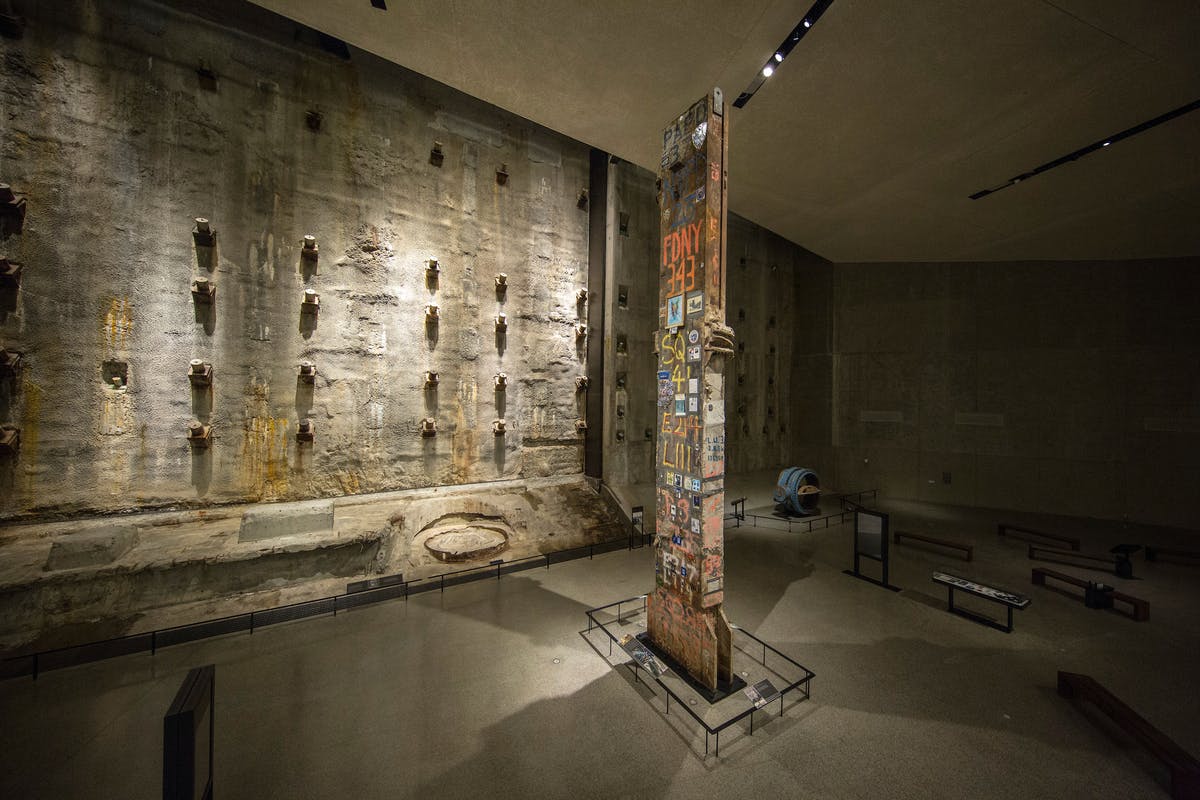 Exposition muséale présentant une grande poutre métallique recouverte de divers autocollants et décalcomanies, entourée d'un mur de béton et de vitrines.