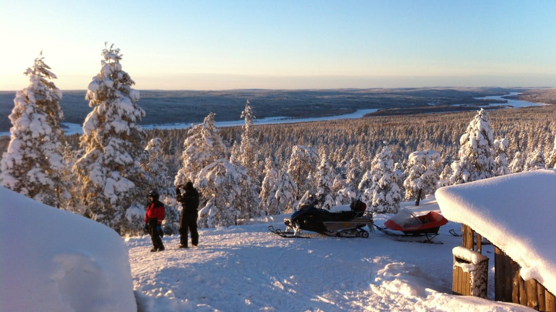 Δύο άνθρωποι με χειμερινά ρούχα στέκονται κοντά σε οχήματα χιονιού σε έναν χιονισμένο λόφο με θέα ένα τεράστιο, χιονισμένο δάσος κάτω από έναν καθαρό ουρανό.