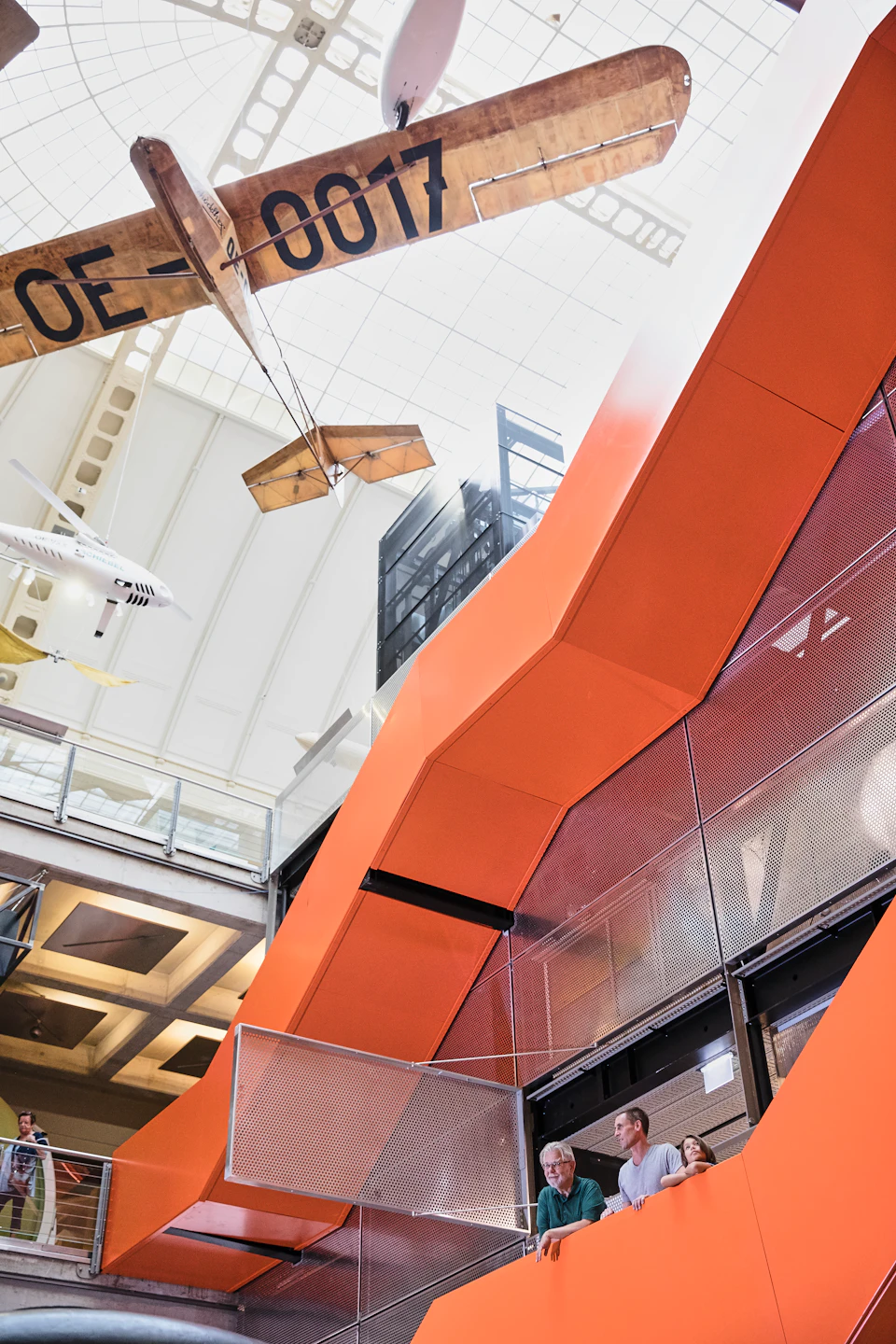 Interior of a modern building with orange structures, hanging airplane models, and a high ceiling with skylights.