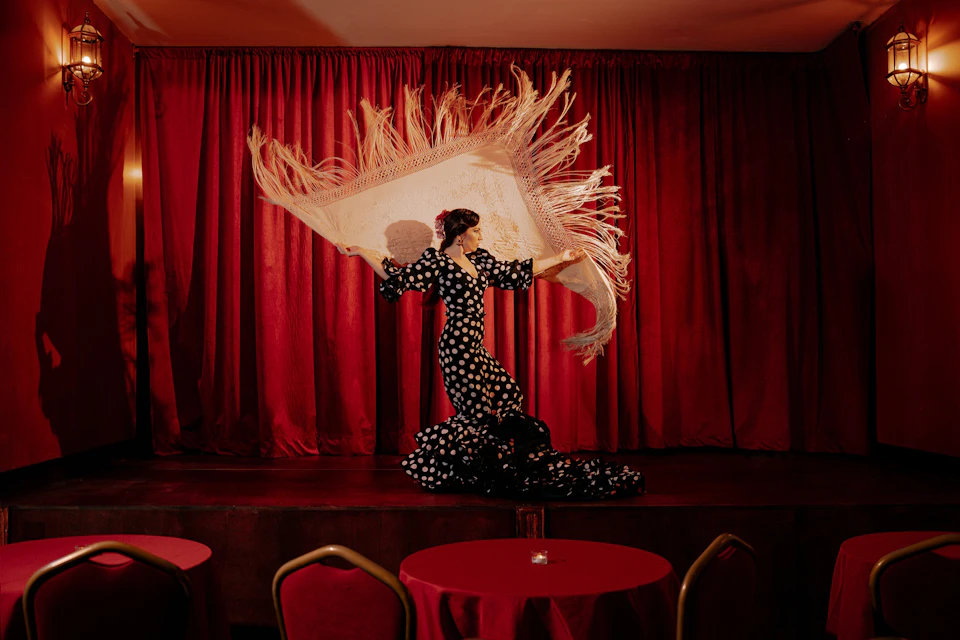 A dancer in a black and white polka-dot dress holds a fringed shawl on a stage with red curtains and chairs in the foreground.