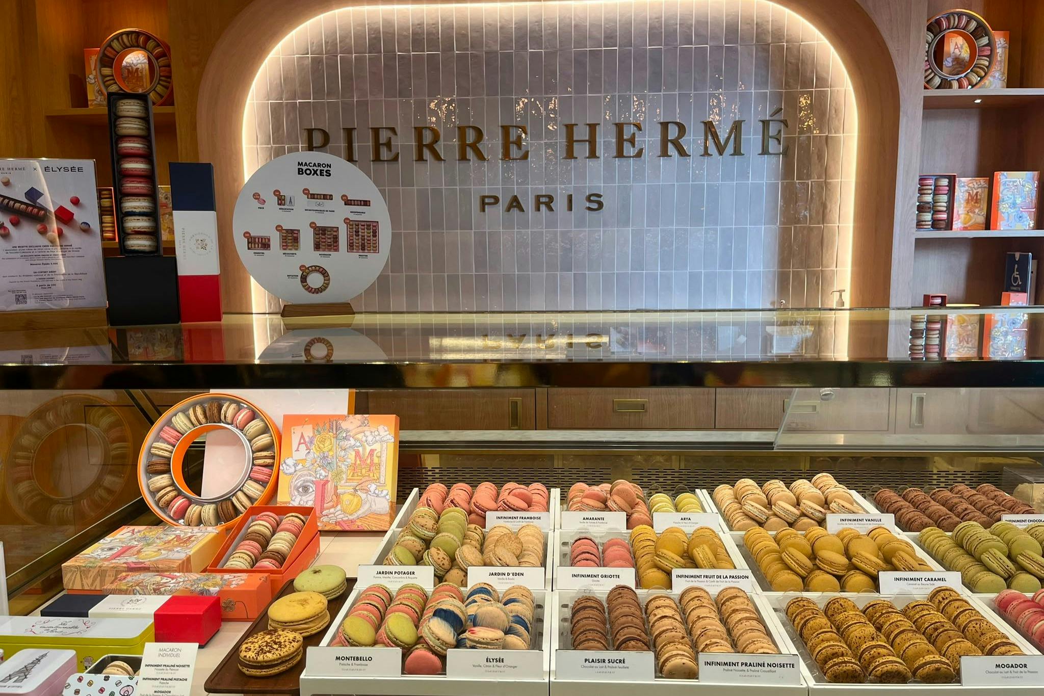 A display of colorful macarons arranged in trays at a Pierre Hermé store, with branded signage in the background.
