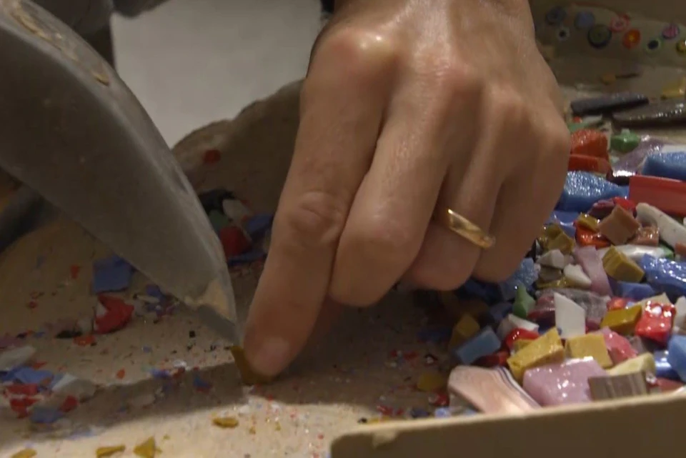A hand holding small colored pieces, likely for crafting, with a knife nearby on a surface containing mosaic tiles.
