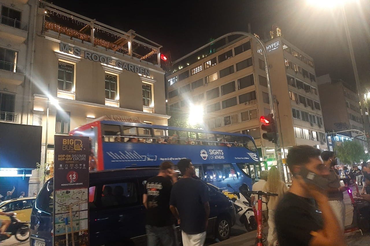 Άνθρωποι που περπατούν κοντά σε ένα διώροφο τουριστικό λεωφορείο τη νύχτα σε μια πόλη. Τα κτίρια και τα φώτα είναι ορατά στο παρασκήνιο.