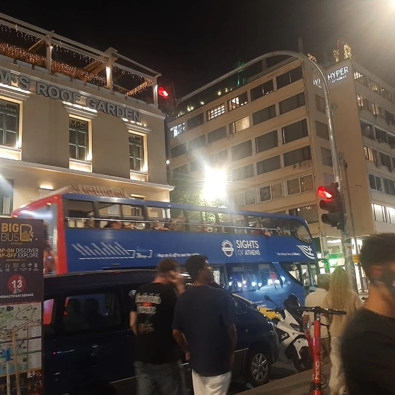 People walking near a double-decker tour bus at night in a city. Buildings and lights are visible in the background.