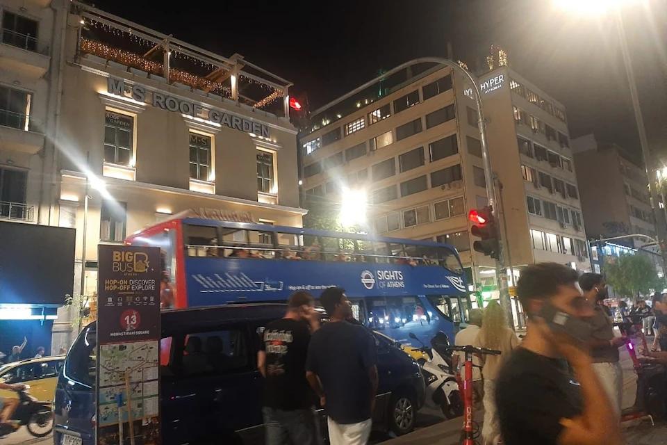 People walking near a double-decker tour bus at night in a city. Buildings and lights are visible in the background.