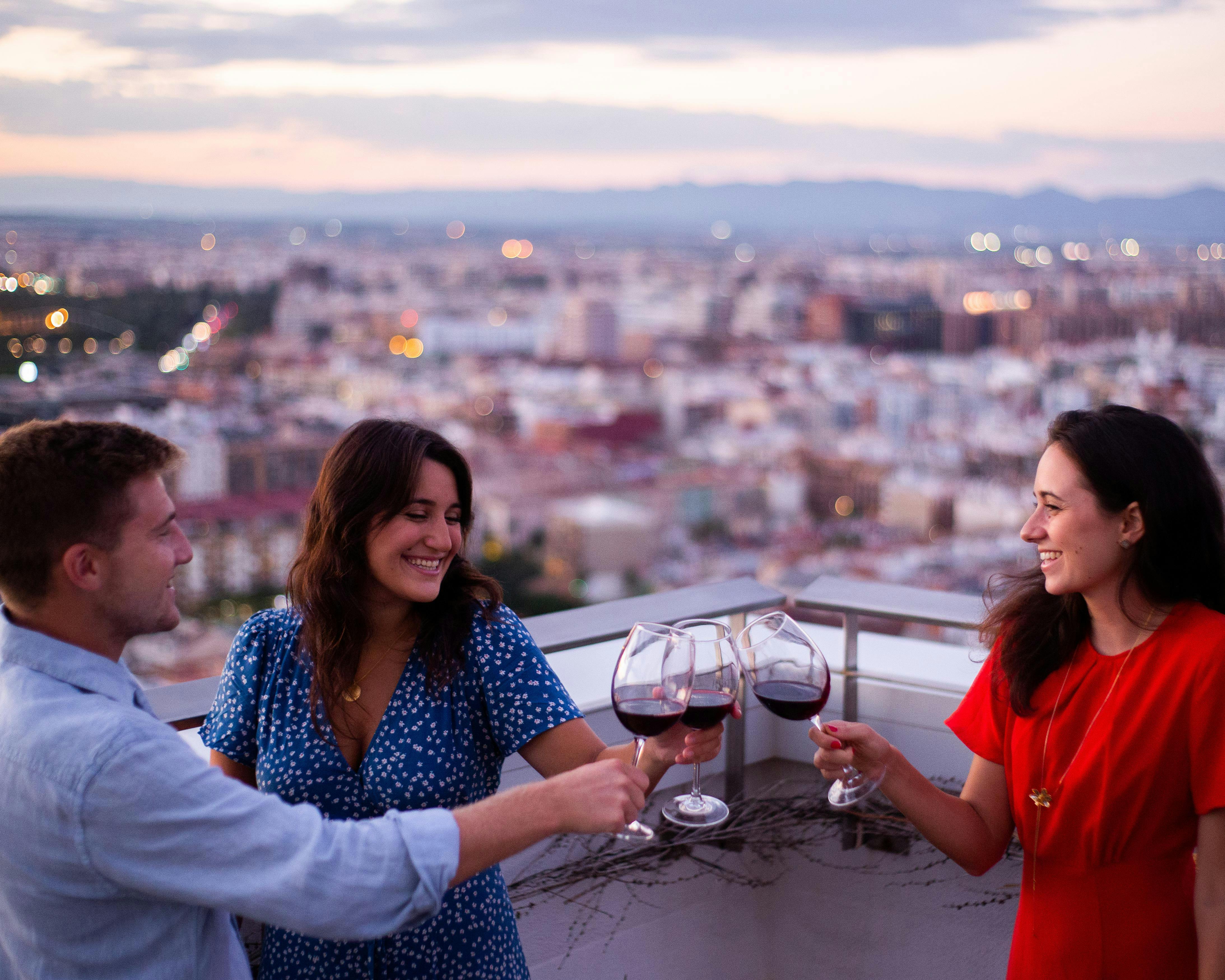 해가 질 무렵 도시 풍경을 배경으로 옥상에서 와인잔을 쥐고 있는 세 사람.