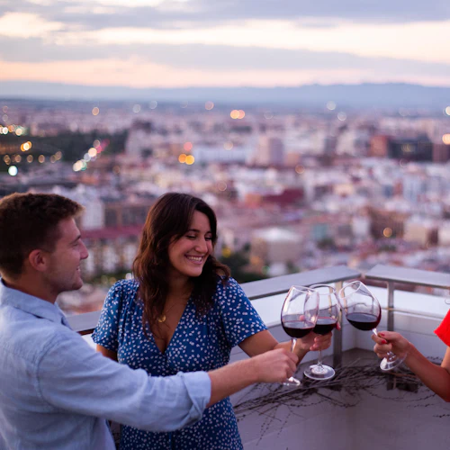 Tre personer klirrar vinglas på en takterrass med en stadsbildsbakgrund under solnedgången.