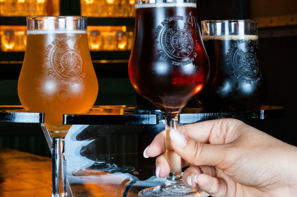 A person holding a glass of dark beer, with two other glasses of beer in the background on a reflective table.