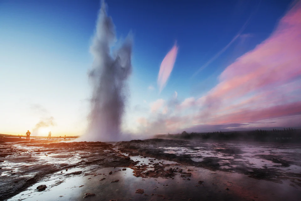 A geyser erupts, shooting water high into a clear, colorful sky at sunset with rocky, wet ground in the foreground.