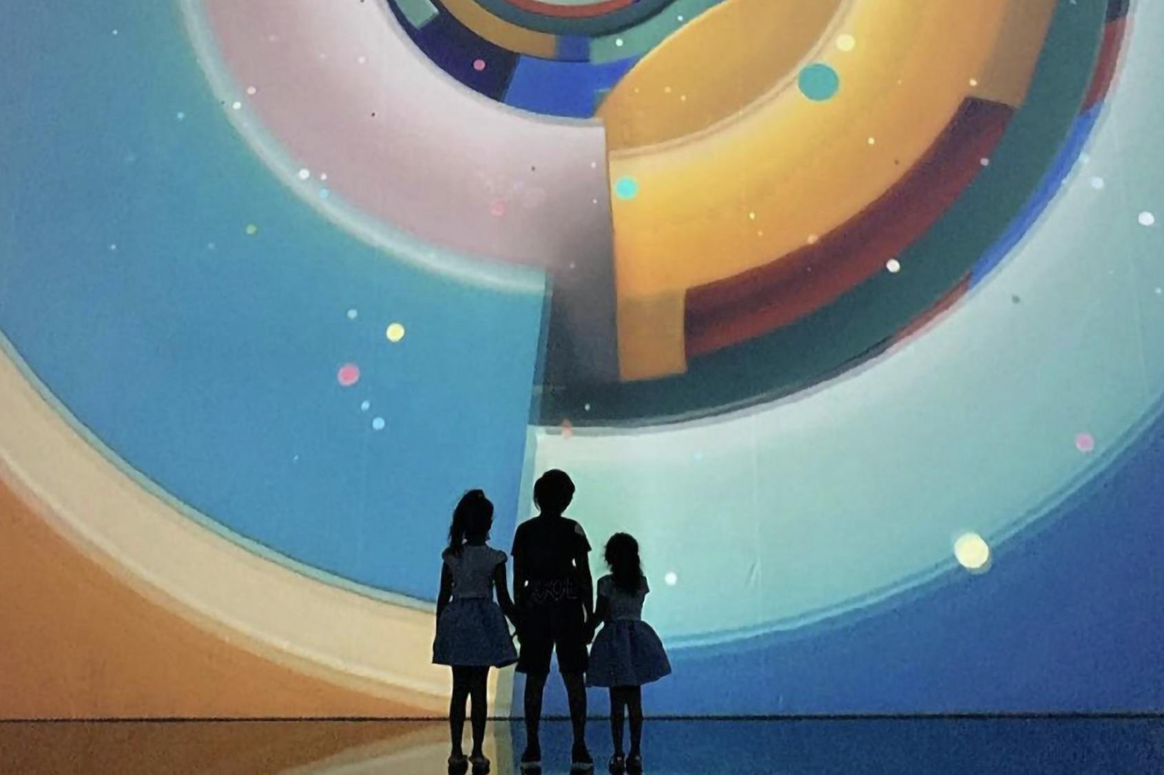 Three children stand in front of a colorful, abstract digital display featuring circular and geometric patterns.