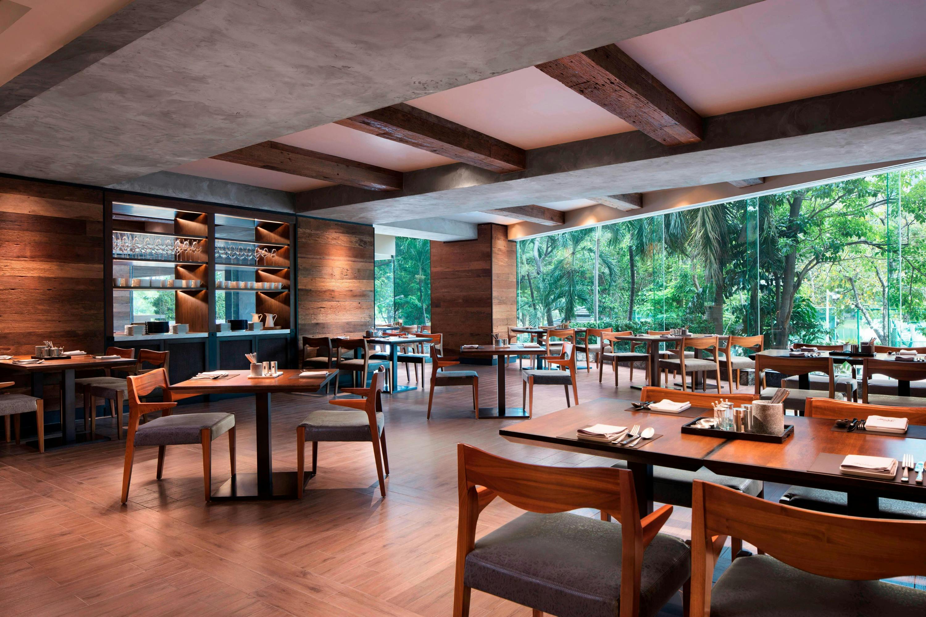 Modern restaurant interior with wooden furniture, large windows overlooking greenery, and a cozy atmosphere.