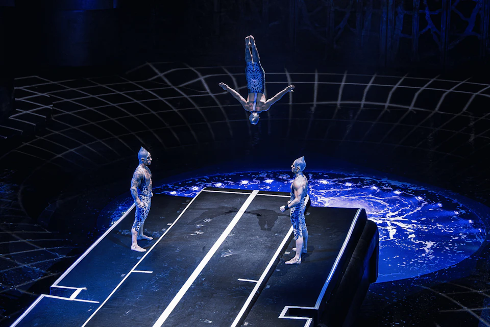 Performers in blue costumes on a stage with a trampoline-like setup; one is mid-flip in the air.