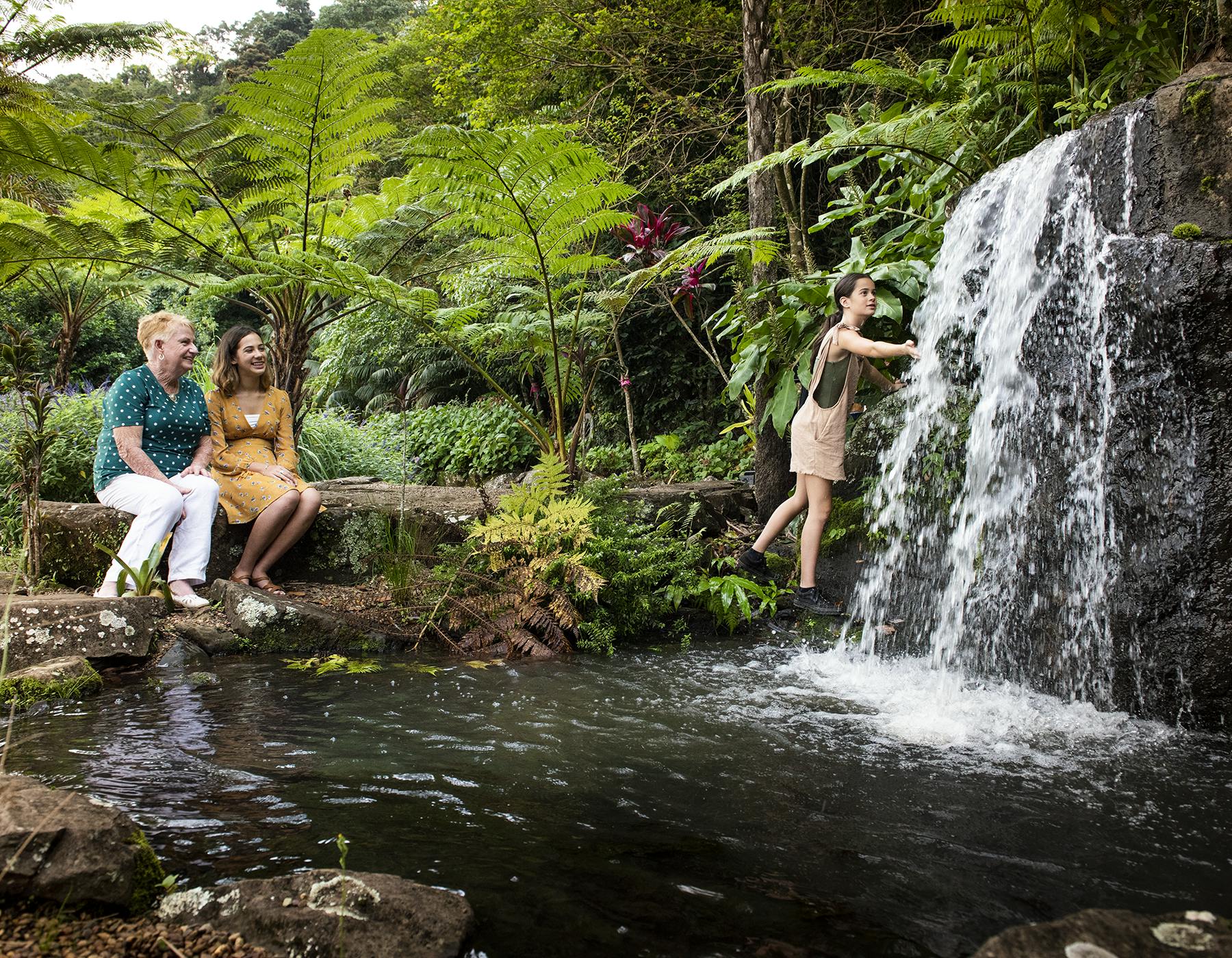 Trzy osoby w bujnym ogrodzie; dwie siedzące nad stawem, uśmiechnięte, a jedna bawiąca się pod wodospadem.