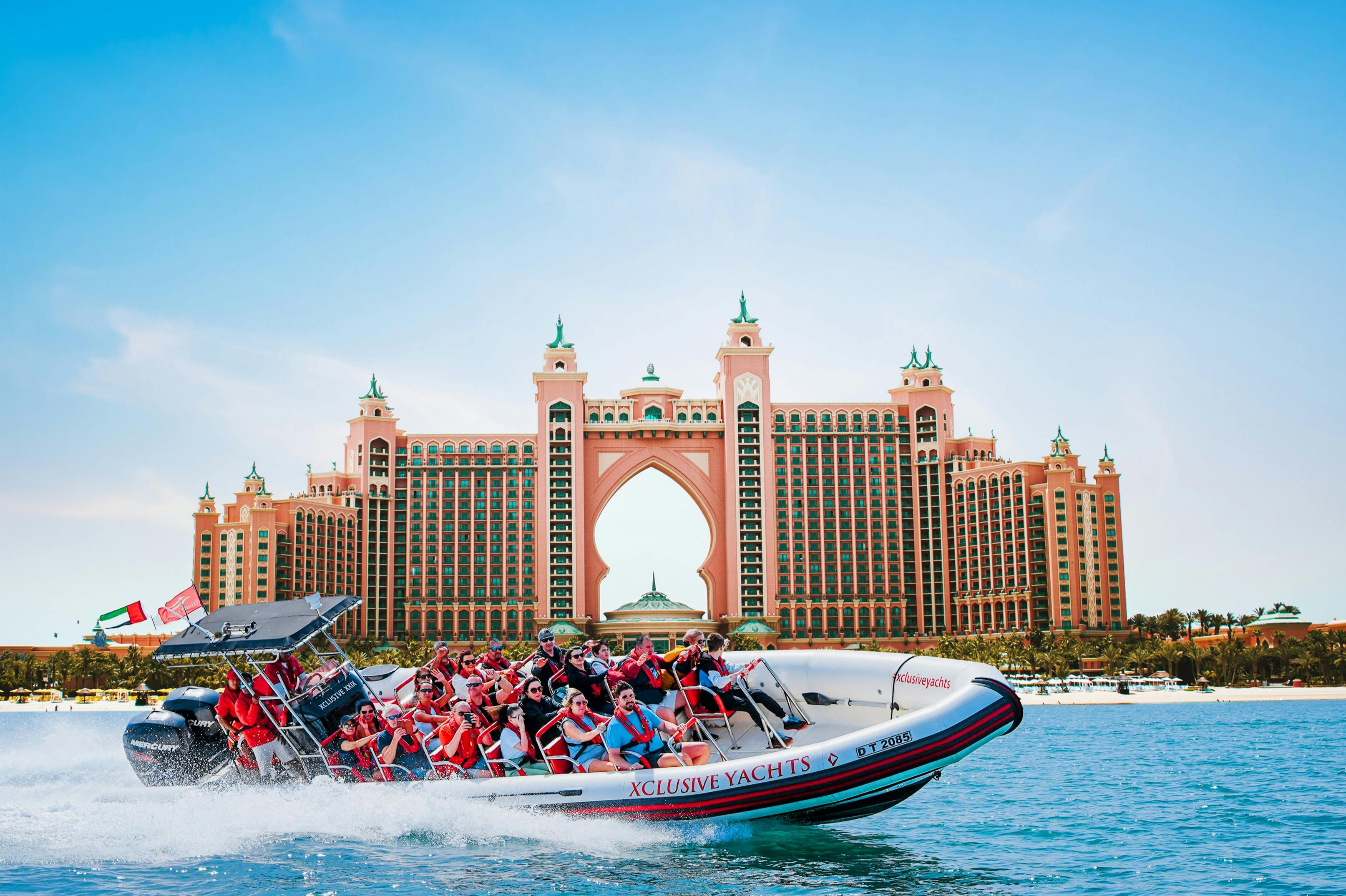 Lidé si užívají jízdu motorovým člunem před velkým růžovým luxusním hotelem ve tvaru oblouku pod jasně modrým nebem.