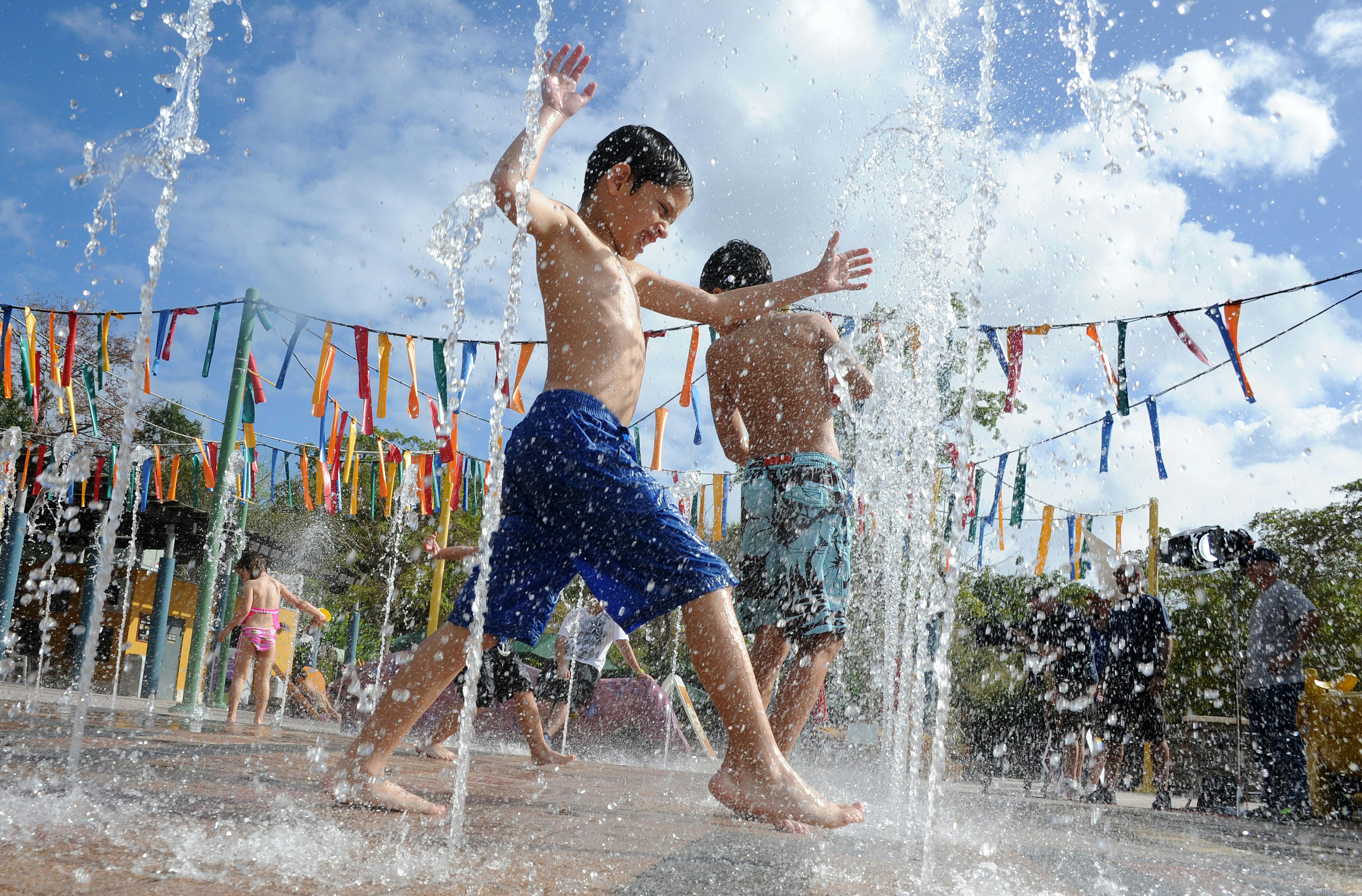Børn leger i et springvand med vandstråler under en solrig himmel, omgivet af farverige dekorationer og bannere.