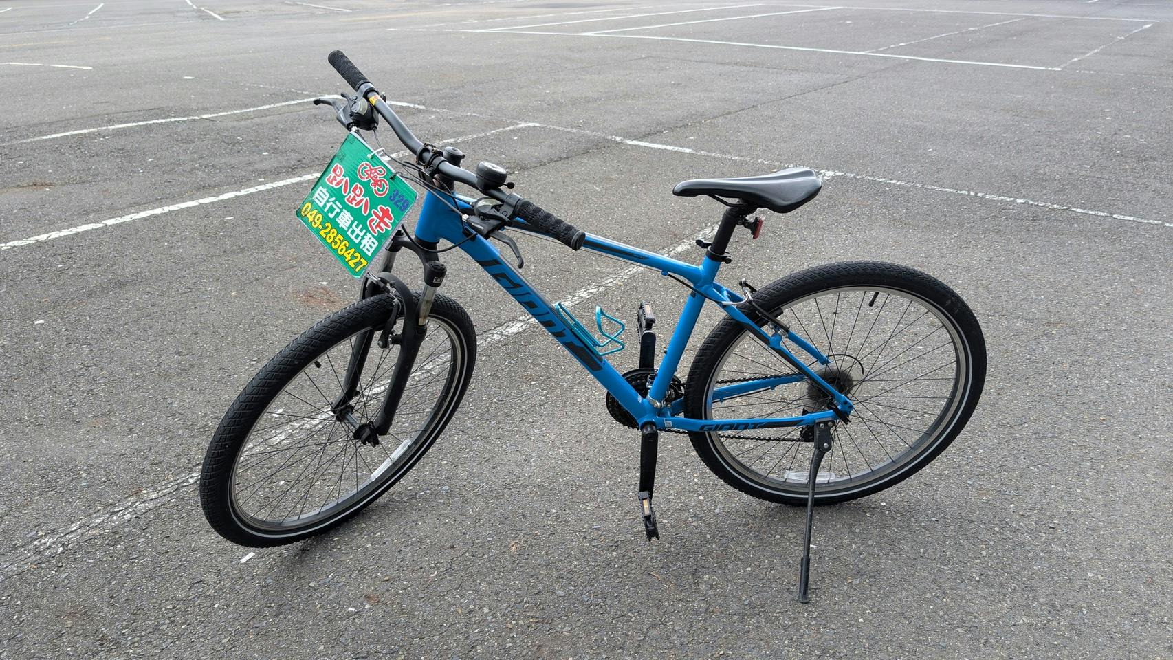 A blue mountain bike parked in an empty parking lot, with a small advertisement sign attached to the handlebars.