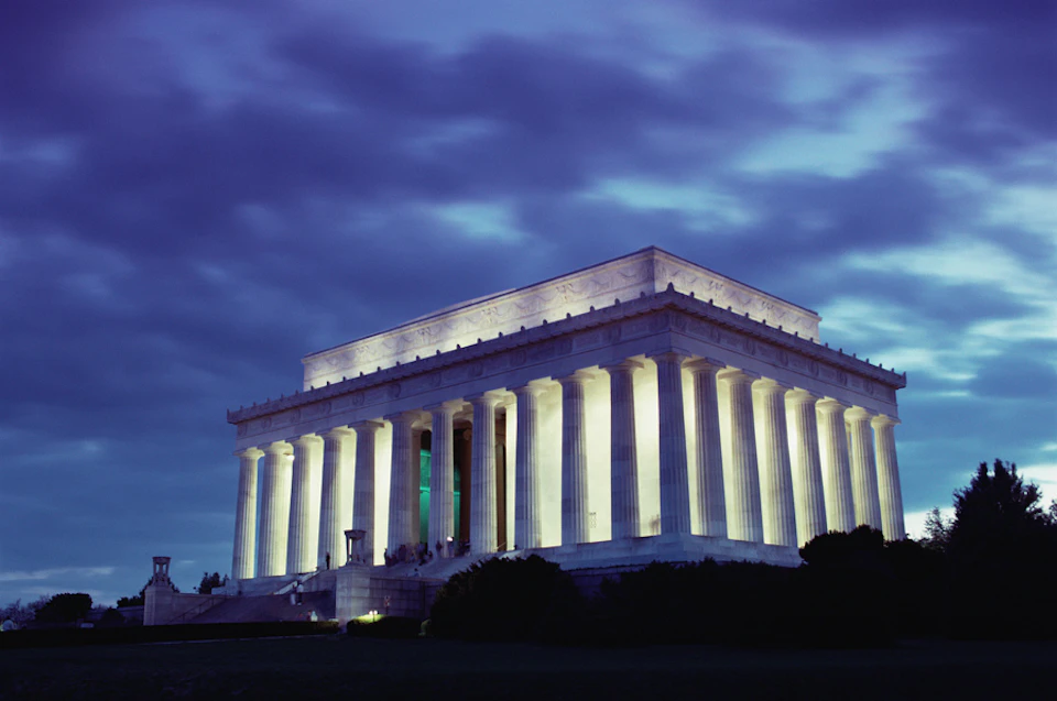 The image shows the Lincoln Memorial illuminated at night, with a cloudy sky in the background.