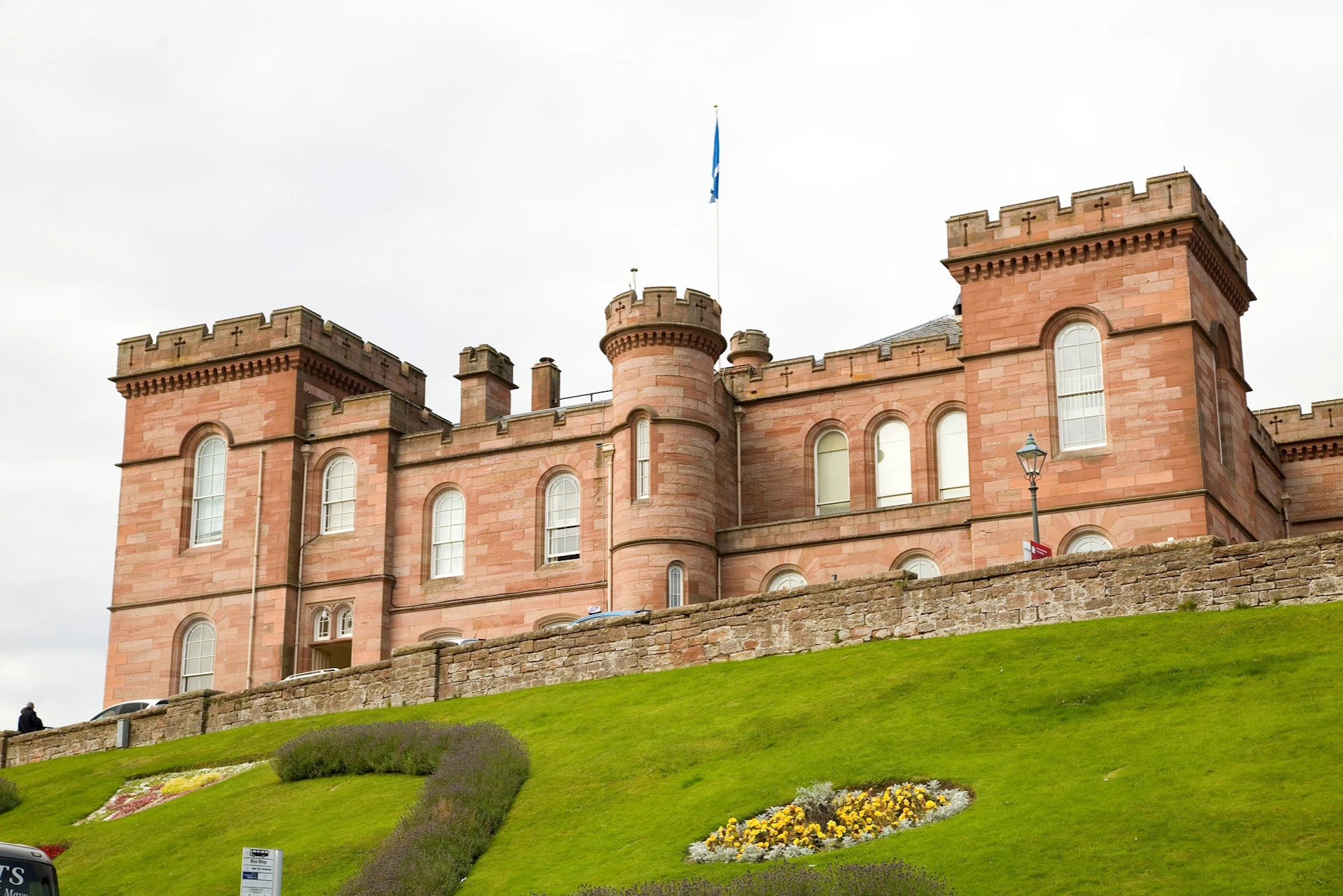 A historic red sandstone castle with a flag on top, surrounded by green lawns and colorful flowers on a cloudy day.