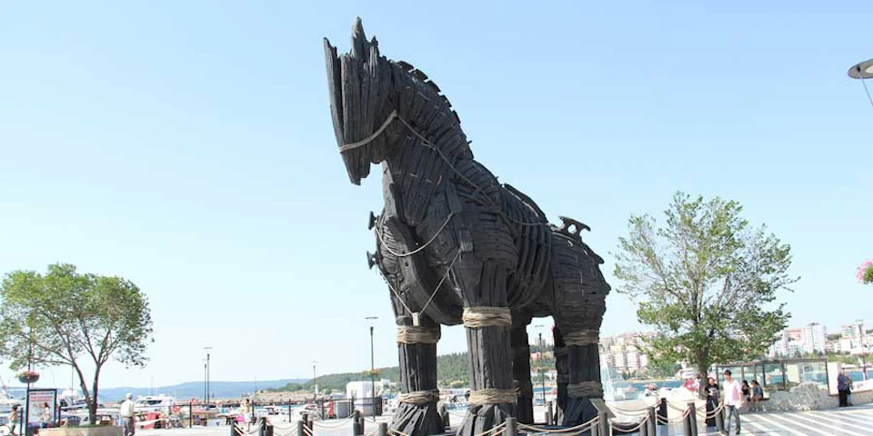 Large wooden horse sculpture with ropes, standing in an outdoor area near a waterfront, under a clear sky.