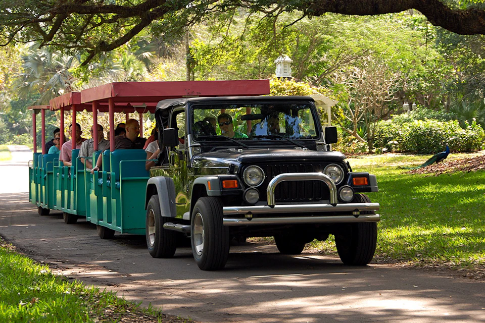 A black Jeep towing a small, covered passenger trailer with people seated, driving along a shaded park path surrounded by greenery.