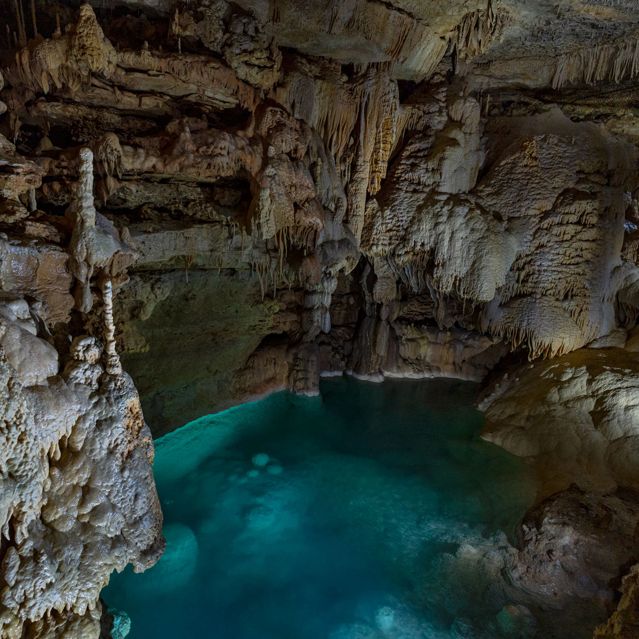 A subterranean cave with teal water, stalactites, and stalagmites, showcasing intricate rock formations in low lighting.