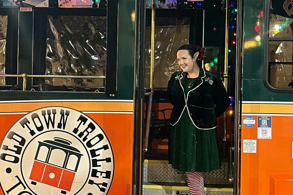 A person dressed in festive clothing stands in the doorway of a trolley decorated with lights.