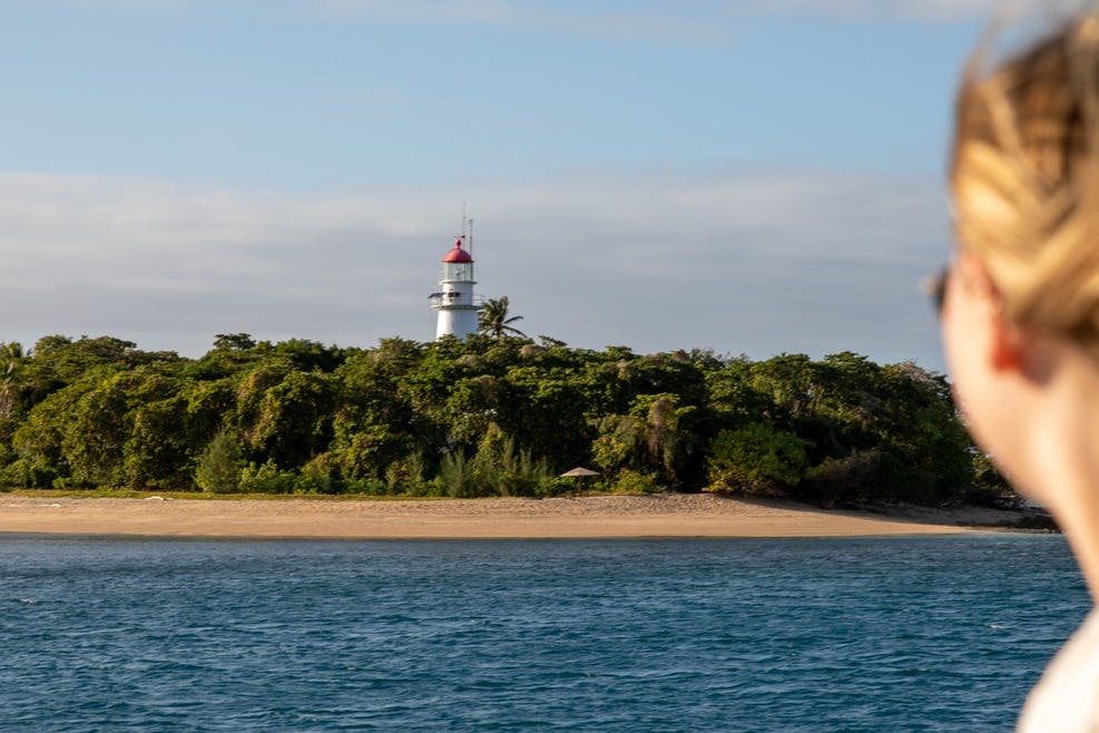 A coastal scene with a red-topped white lighthouse behind dense greenery and a sandy beach, viewed from the water.