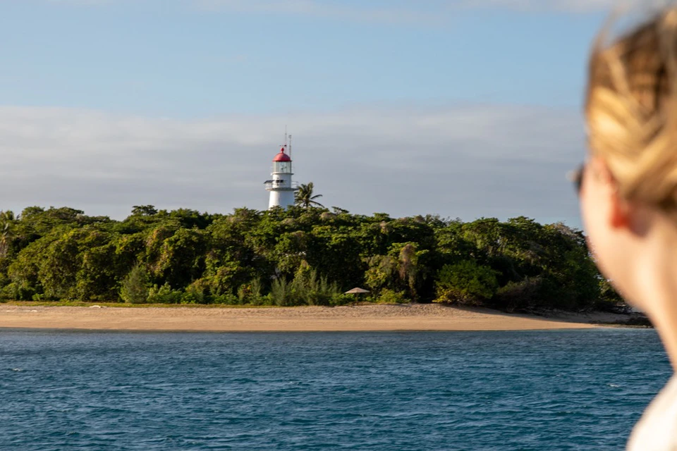 A coastal scene with a red-topped white lighthouse behind dense greenery and a sandy beach, viewed from the water.