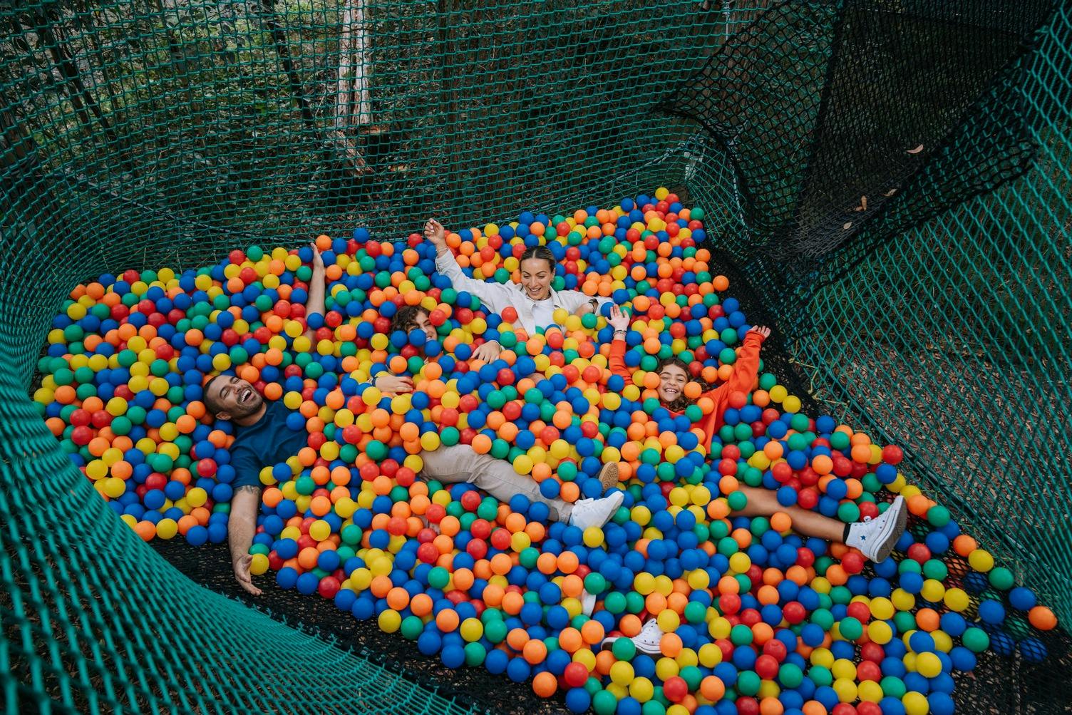 四个人躺在一个五彩缤纷的球坑里，周围是户外绿网，微笑着享受这一刻。
