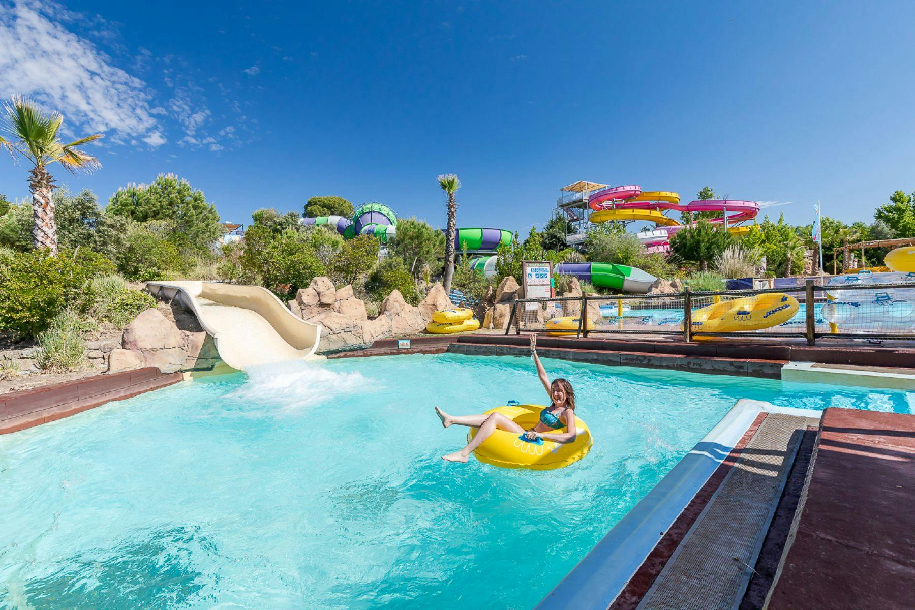 Une personne sur une bouée jaune dans la piscine d'un parc aquatique, avec des toboggans et de la végétation en arrière-plan, sous un ciel bleu.