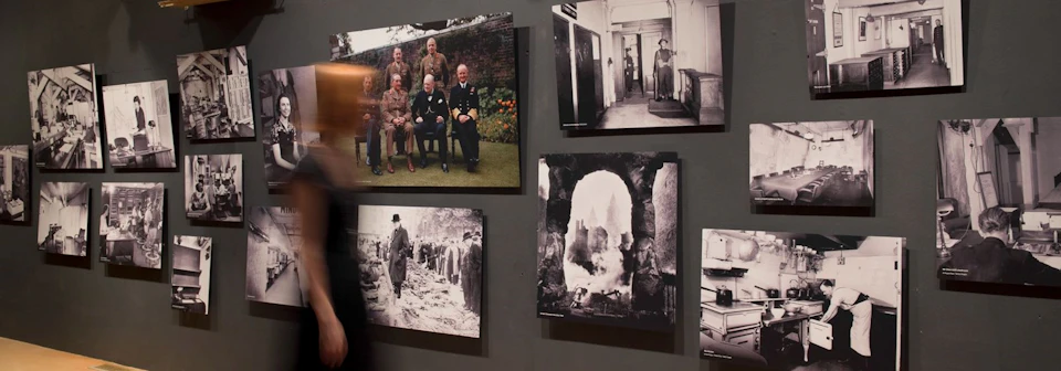 A wall displays five historical photos, including group portraits, scenes of destruction, and a man walking through ruins.