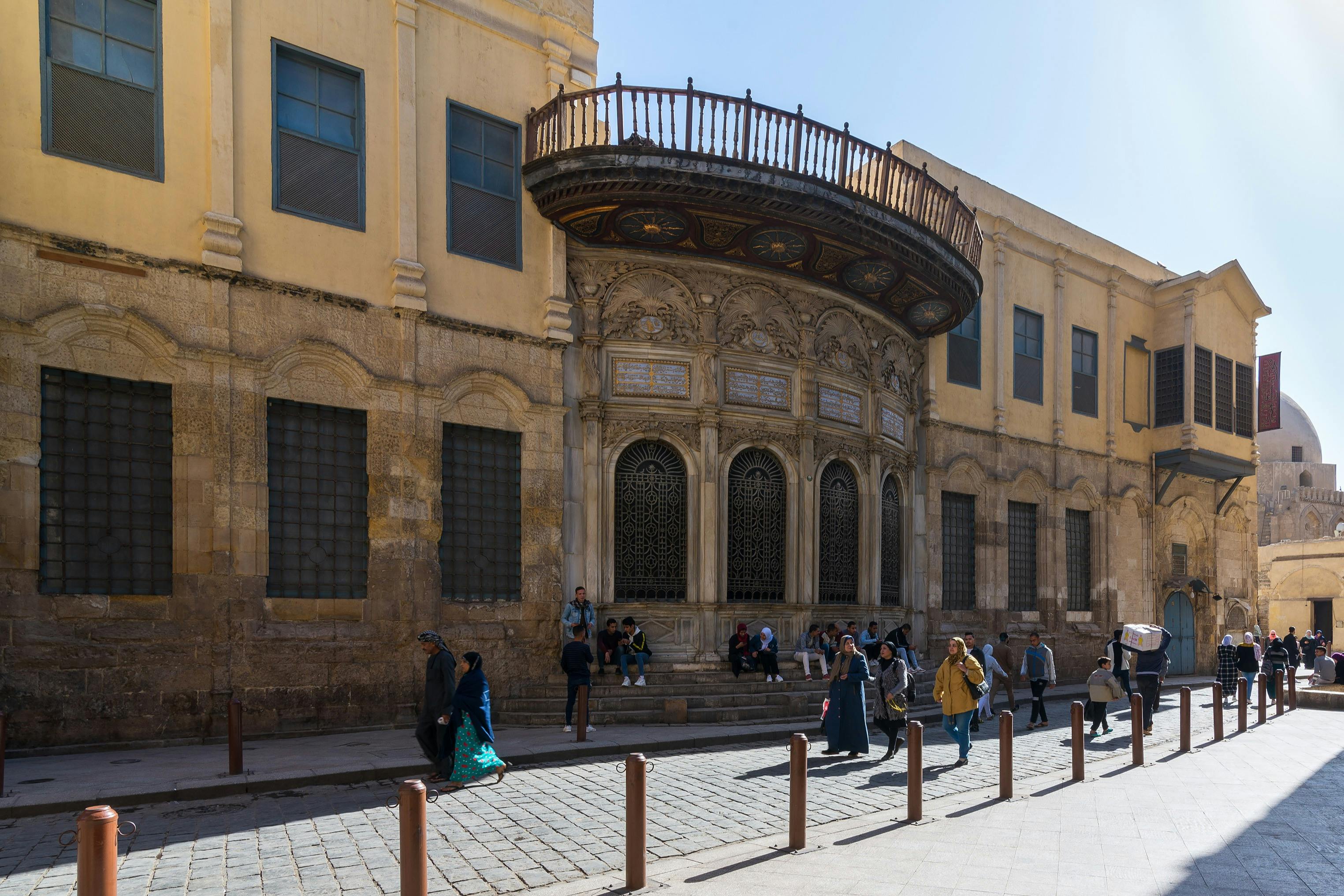 Люди гуляют по мощеной улице перед старым зданием с богато украшенными арками и большим декоративным балконом.