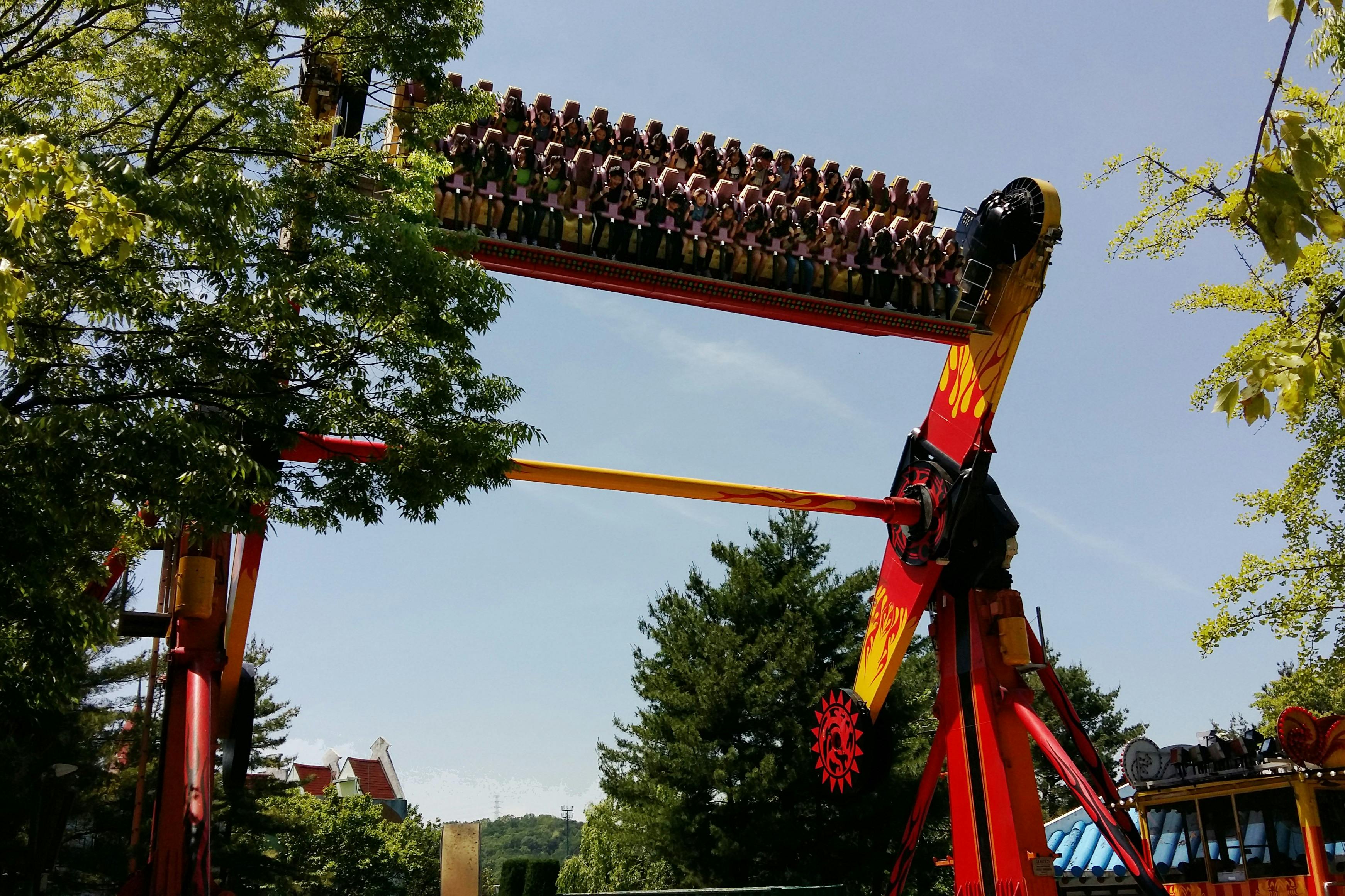 Persone sedute su un'altalena di un parco divertimenti, sollevate a testa in giù su uno sfondo di alberi e cielo sereno.