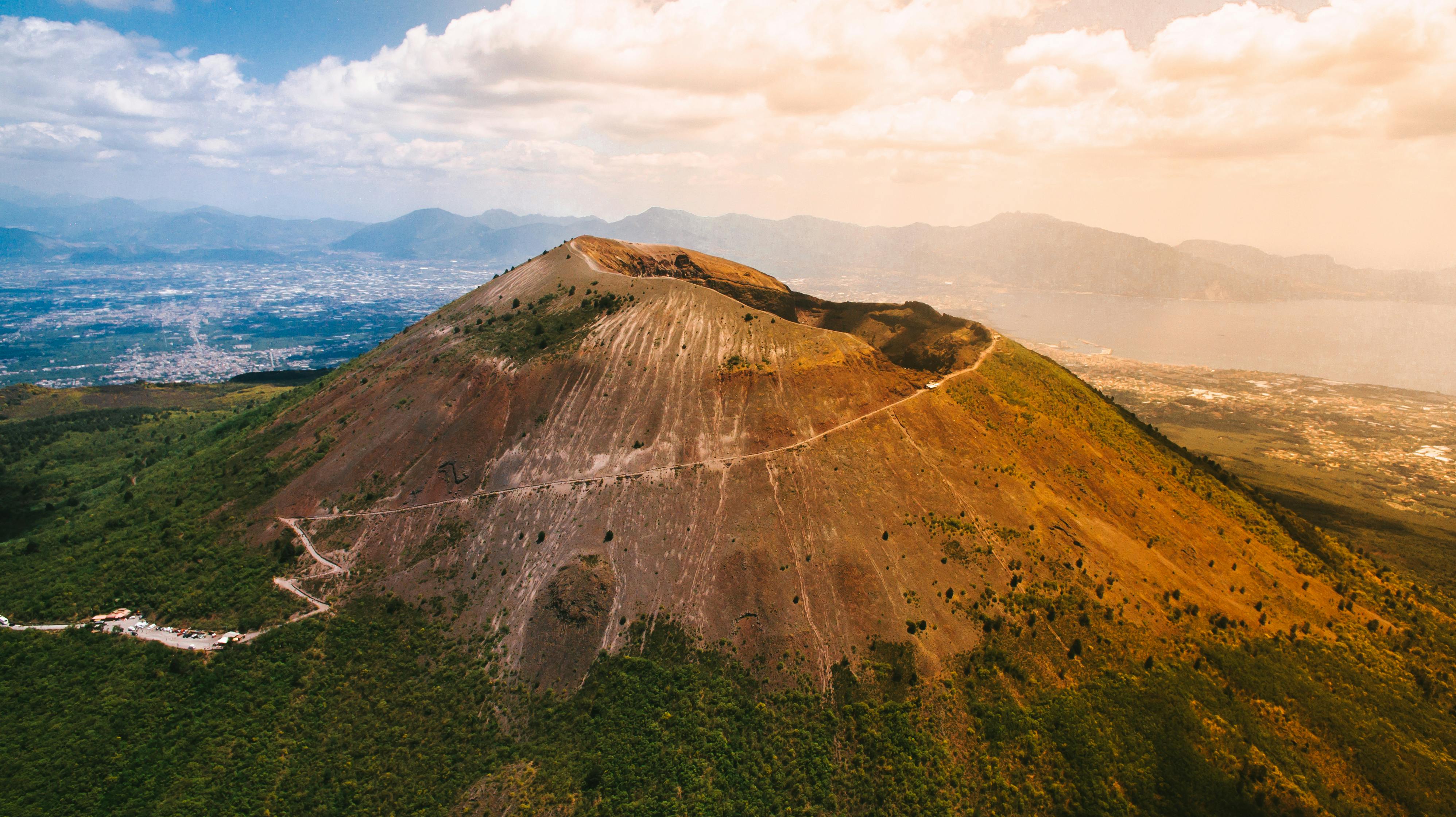 Aerial view of a mountain with a winding path, surrounded by green vegetation, and a cityscape and bay in the background under a cloudy sky.