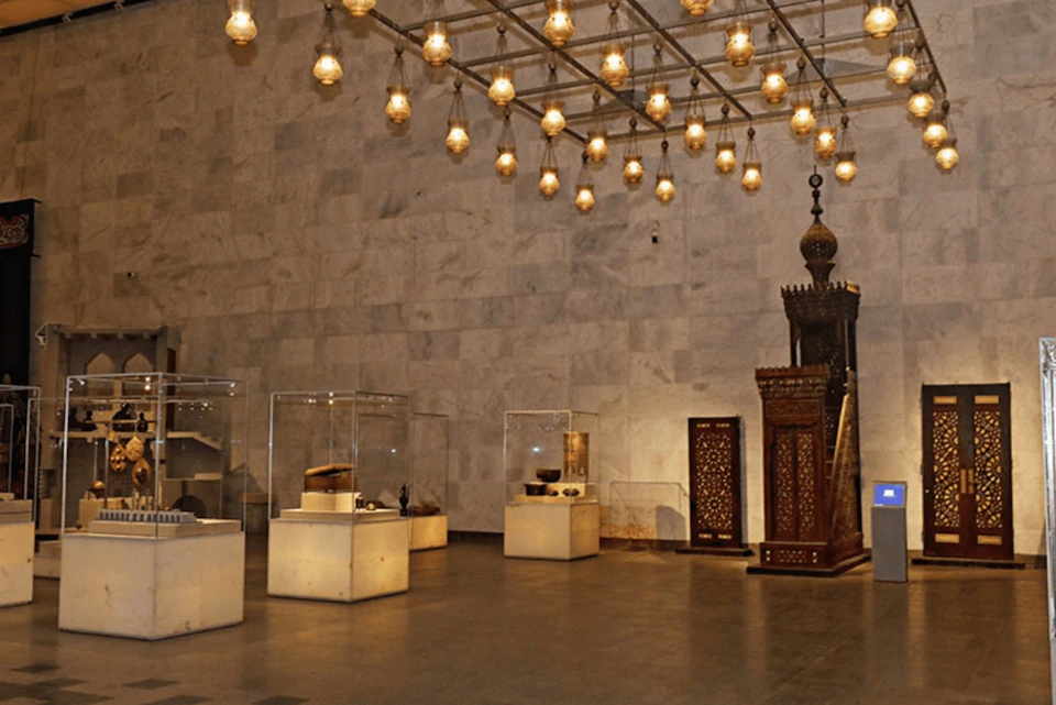 A museum display area with artifacts in glass cases and ornate wood carvings, illuminated by numerous hanging lamps.