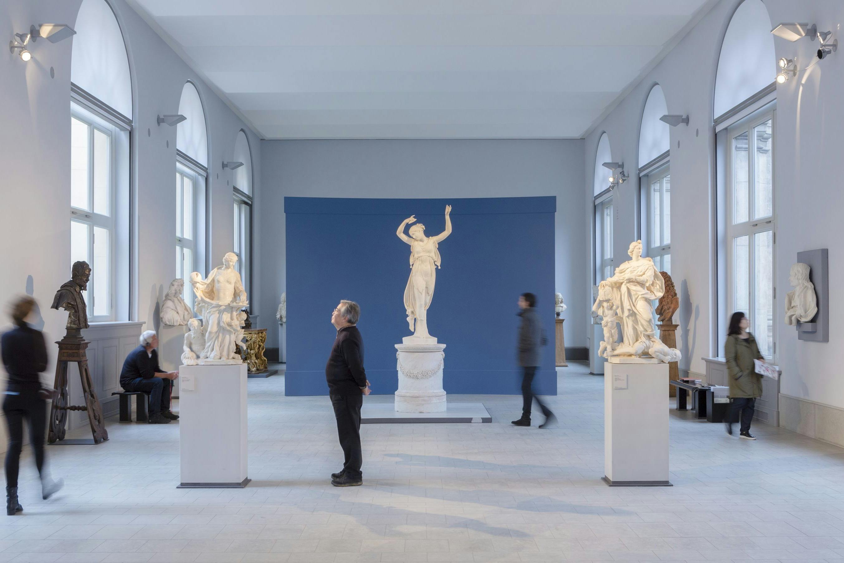 An art gallery with sculptures on display, including a central white statue on a pedestal, and people observing the exhibits.