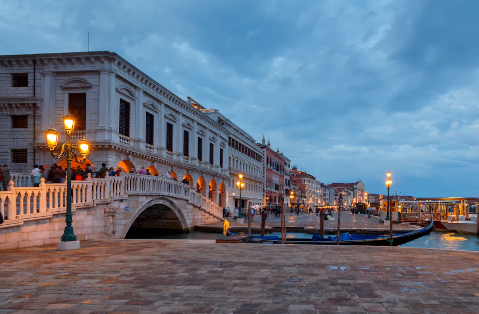 Evening view of a canal in Venice with a stone bridge, lit street lamps, and historic buildings. People are walking along the promenade.