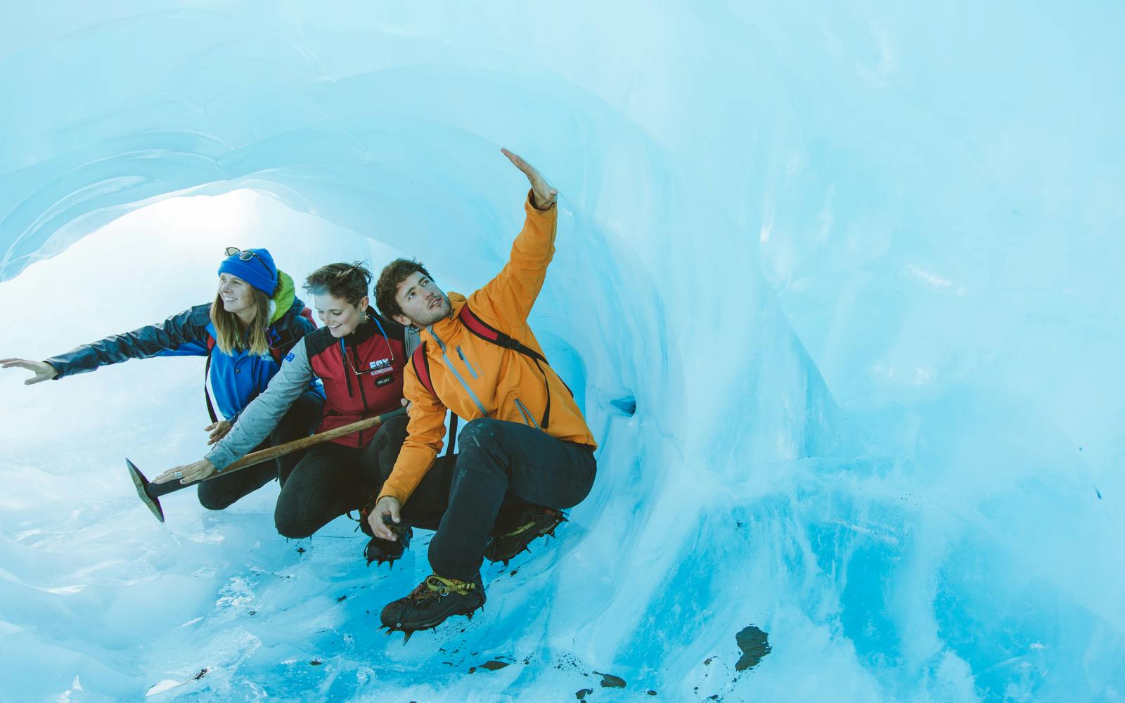 Three people in warm clothing exploring an ice cave, with one gesturing towards the ceiling.