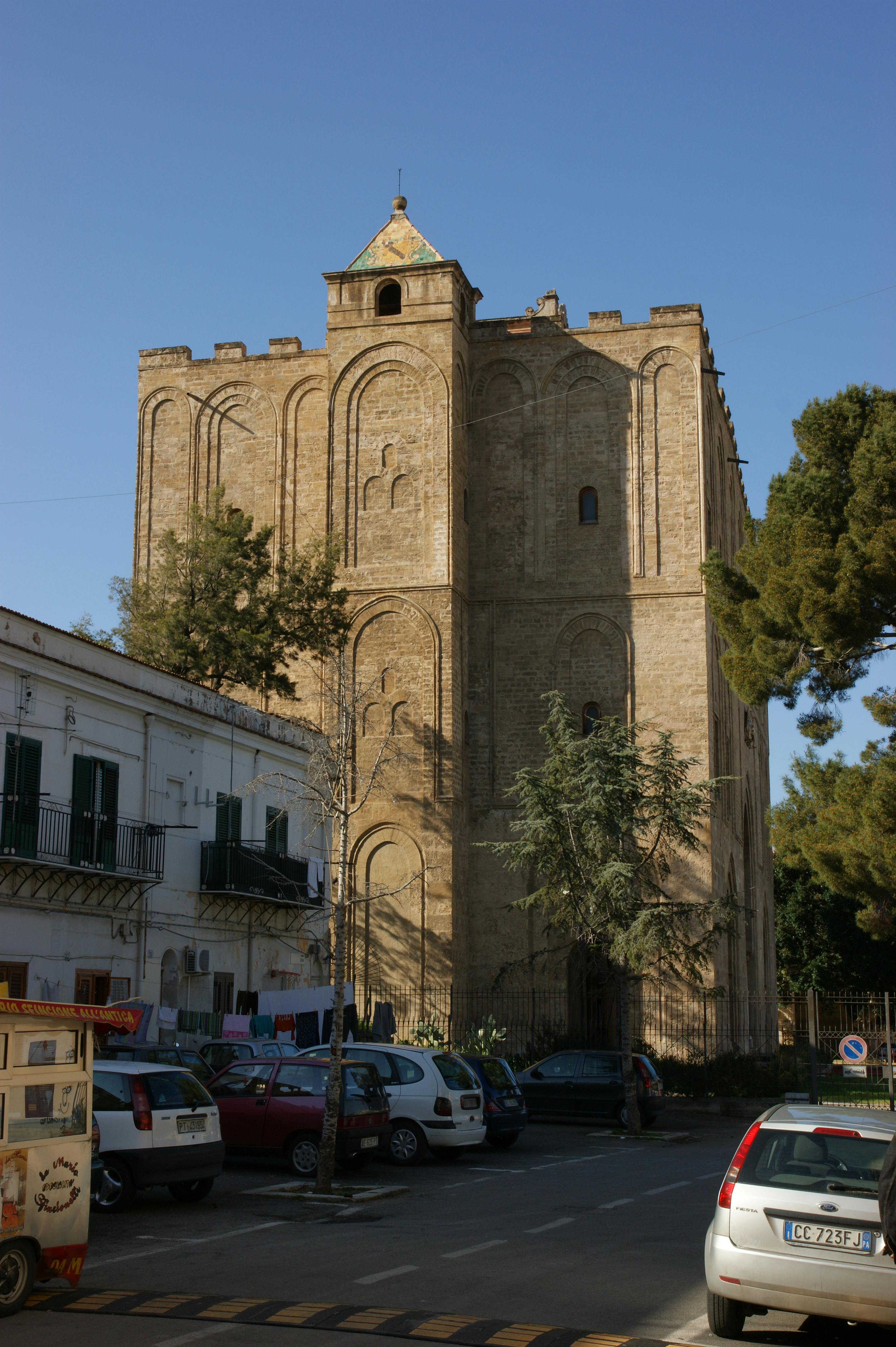 Tall, rectangular stone building with arched details, surrounded by parked cars, trees, and an adjacent white residential building.