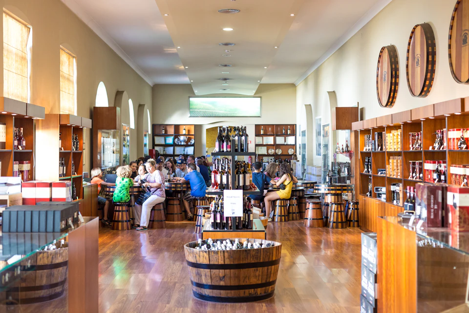 People seated at tables made of barrels in a brightly lit room with wooden floors, shelves of bottles, and arched windows.
