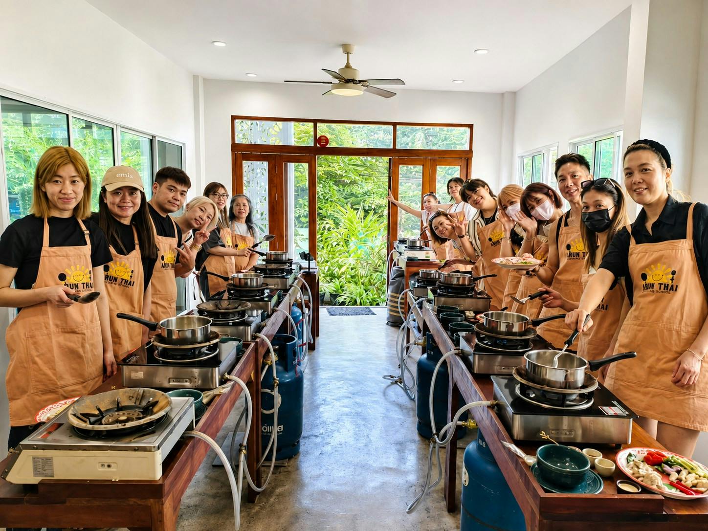 A group of people wearing aprons in a cooking class setup with stoves and pans, inside a room with large windows and greenery outside.