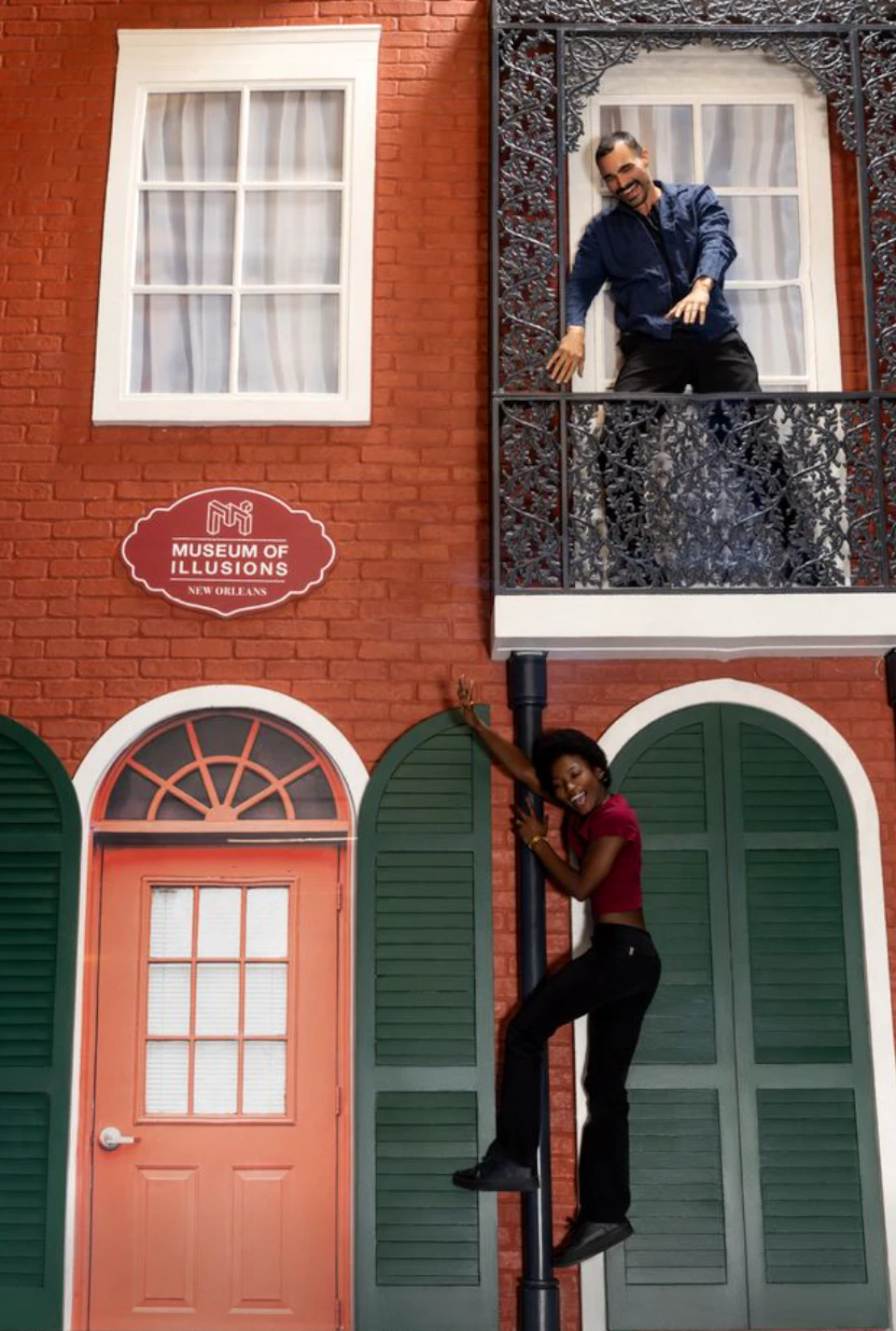 A woman poses as if climbing a pole in front of a building with a sign reading 'Museum of Illusions New Orleans,' while a man leans from a balcony above.