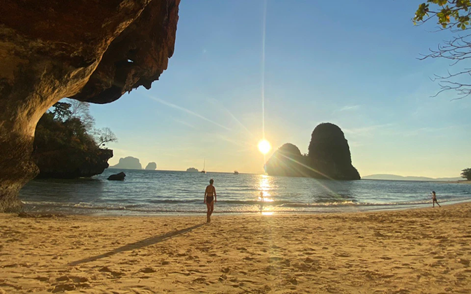 A person in swimwear walks on a sandy beach toward the ocean, with large rock formations and the sun setting in the background.