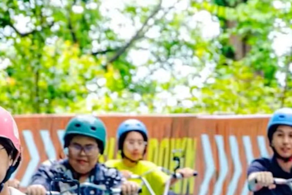 People riding colorful go-karts on a blue track surrounded by greenery, with one person standing nearby.