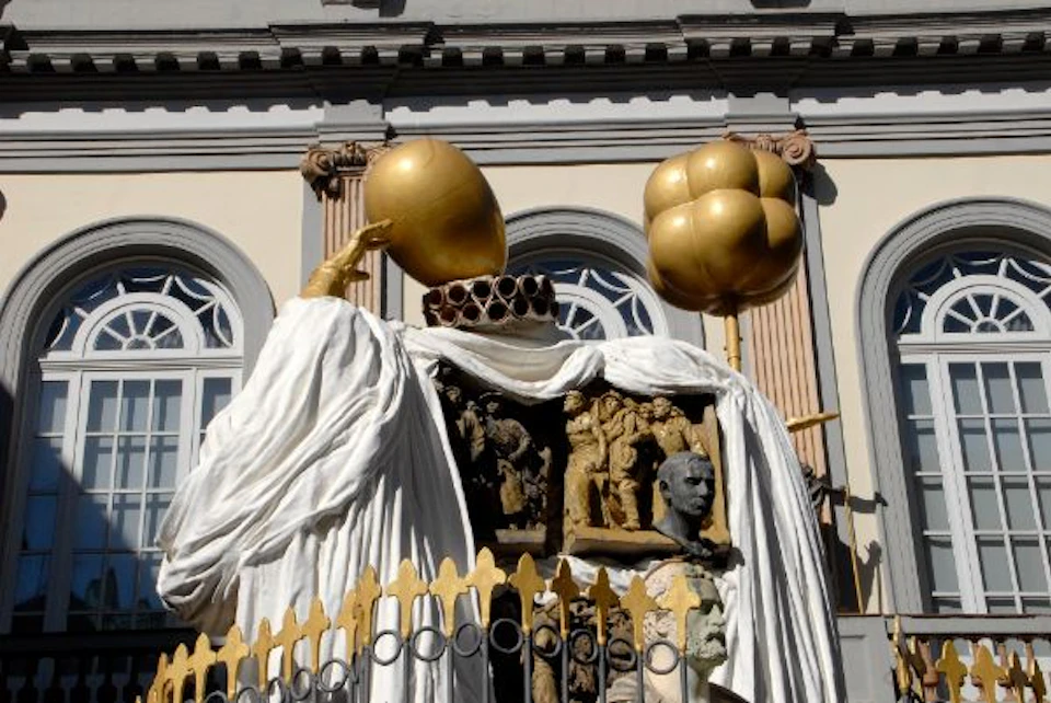Sculpture with golden spheres, draped in white cloth, featuring human figurines and a bust. Set against a building with arched windows.