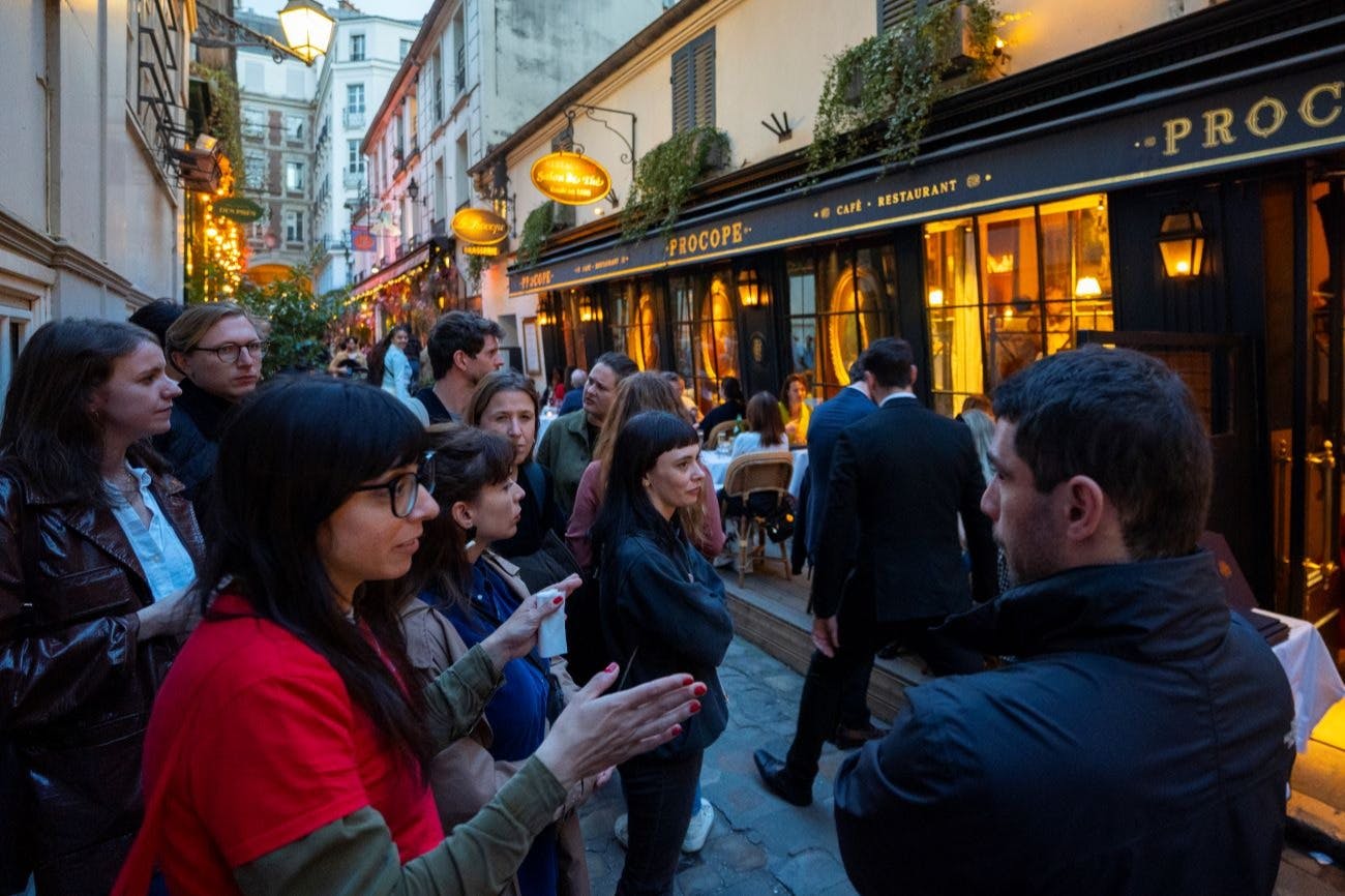 야외 좌석이 마련된 따뜻한 조명의 카페 바깥 자갈길에 한 무리의 사람들이 서 있습니다.