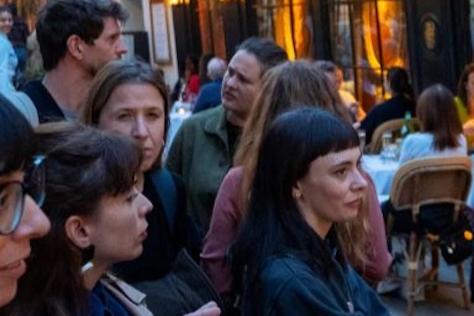 A group of people standing on a cobblestone street outside a warmly lit café with outdoor seating.
